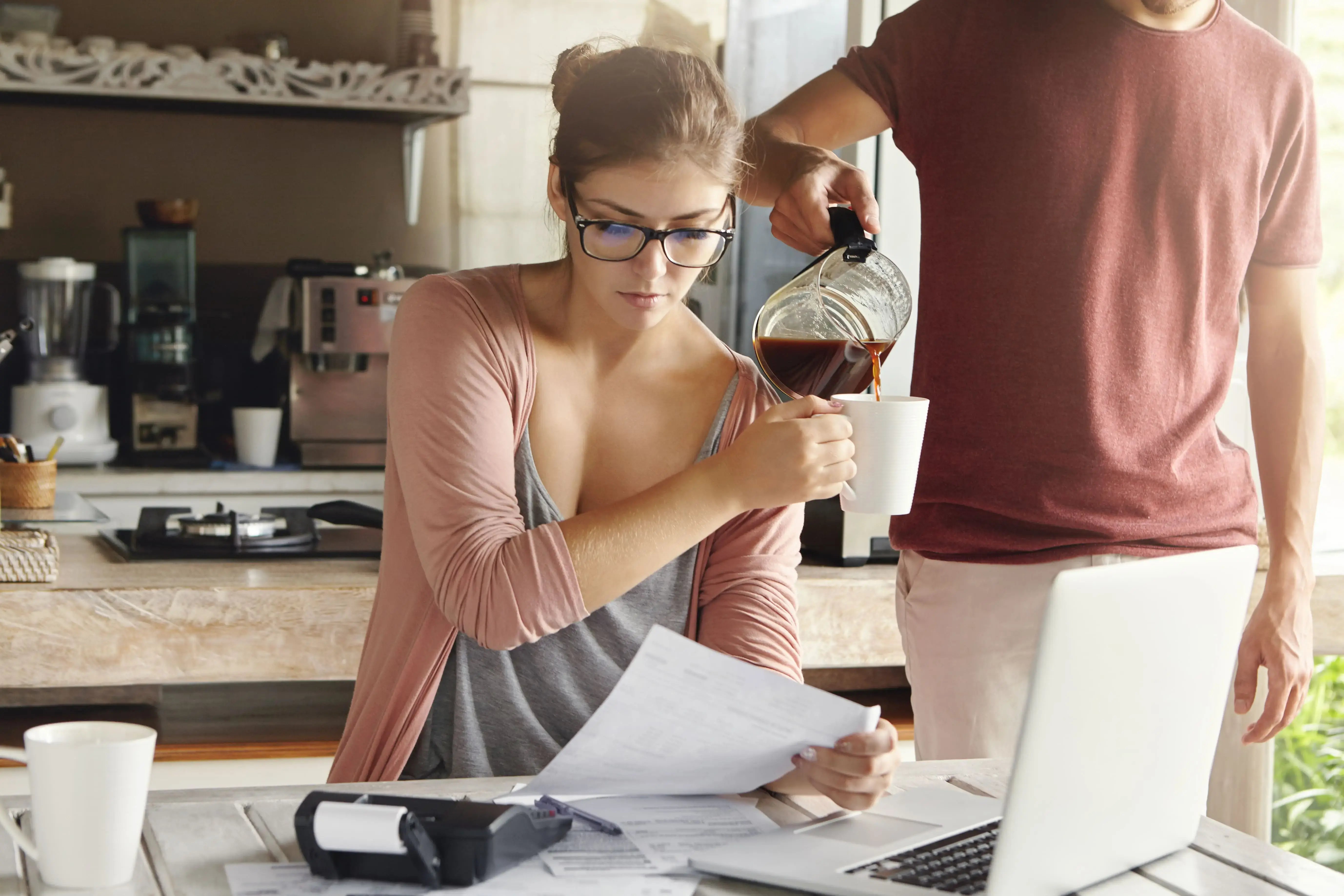 beautiful-woman-glasses-holding-piece-paper-doing-paperwork-paying-taxes-kitchen-table-with-laptop-pc-calculator-it-her-husband-standing-beside-her-adding-coffee-her-mug